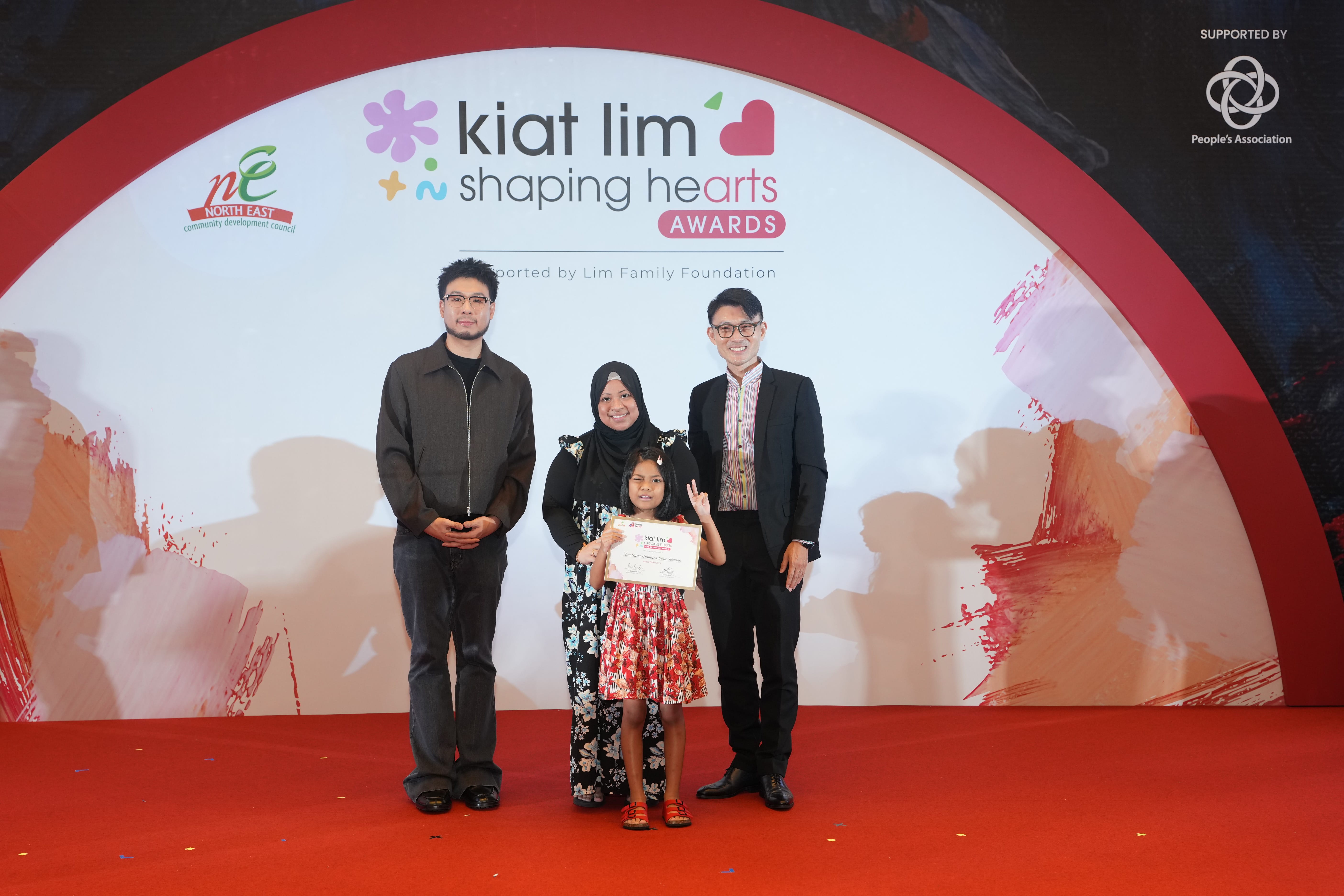 Award recipient standing on stage with the presenter, holding their certificate and posing for a group photo during the award ceremony, with a backdrop reading ‘Kiat Lim-Shaping Hearts Awards'.